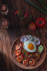 Top-down view of a plate of fried rice with shrimp, sunny side egg, crackers, cucumber, and chopped chili, surrounded by tomatoes, chili peppers, and an egg on rustic wood background