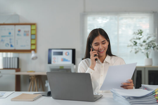 Young asian businesswoman smiling and talking on mobile phone while reviewing documents at her desk in a modern office, showcasing efficiency and multitasking in a professional environment