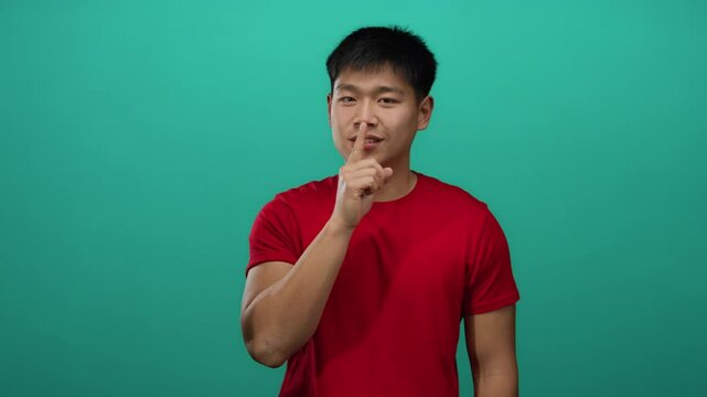 Young man gestures silence with finger on lips against isolated green background wearing red shirt