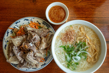 A bowl of pho with beef brisket is paired with a broken rice plate and vegetables on a wood table, captured May 31, 2019, in Seattle, WA, USA