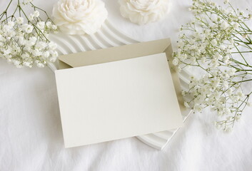 Blank paper card mockup, envelope, white flowers and white table decor candles flowers on white linen textured background. Copy space.Wedding invitation mockup, greeting card, holiday concept.