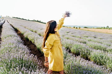 woman in yellow dress stands in lavender field, gently holding bouquet of lavender flowers. She appears serene and contemplative, enjoying beauty and fragrance of blossoms around her.