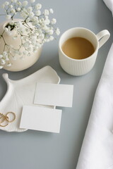 Blank paper sheet cards on home office desk workspace with coffee cup, vase, home decor on grey background. Wedding invitation cards. Flat lay, top view mockup with empty copy space.Life style concept