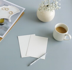 Blank paper sheet cards on home office desk workspace with coffee cup, vase, home decor on blue grey background. Wedding invitation cards. Flat lay, top view mockup with empty copy space.