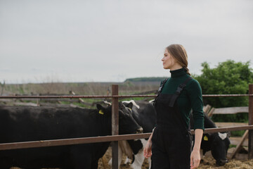 Young female farm worker near a paddock with cows outdoors.