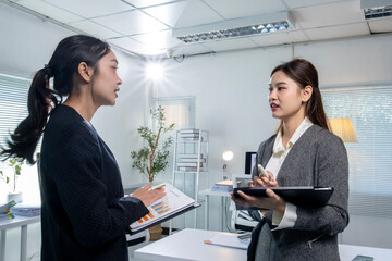 Two women in business suits are talking in a room with a potted plant and a desk