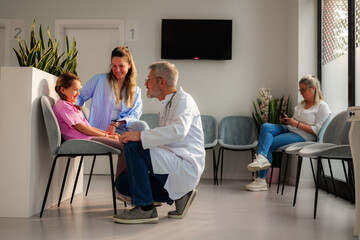 Senior male doctor engaging in conversation with a smiling girl and her mother, while another patient is using a phone in the bright, modern waiting room of a medical clinic