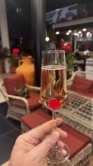 Person’s viewpoint holding champagne glass with cherry at bottom, surrounded by warm bar interior and ambient lighting..