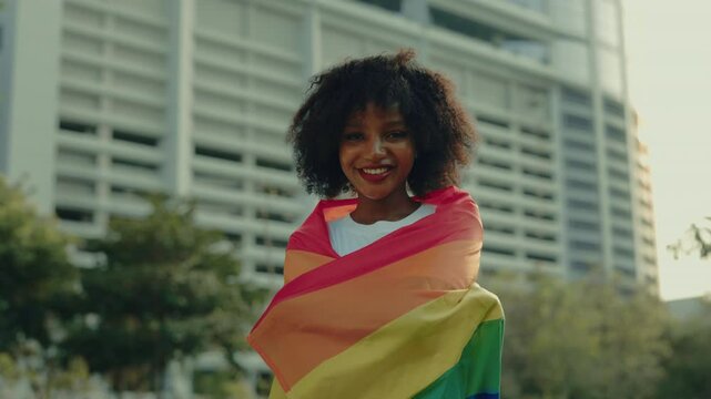 Portrait of pretty African American woman wrapping with pride flag standing near office building. Joyful lady shows support to LGBT community in modern city. Pride month