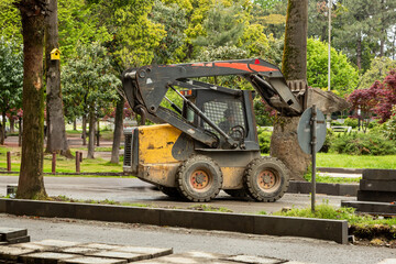 A skid steer loader operating on a park path surrounded by trees during landscaping work