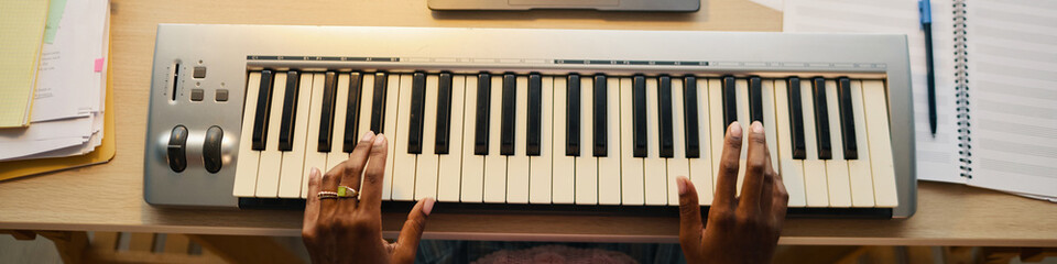 Hands seen playing electronic keyboard in workspace with sheet music and papers surrounding instrument. Overhead view capturing creative process and musical expression