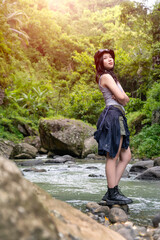 Young Asian Woman Standing on Rocky Riverbank in Forest During Outdoor Adventure