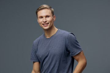 Obraz premium Smiling young man wearing casual blue t-shirt against plain gray background, looking at camera, happy expression, pose indoors, natural lighting, portrait shot, youthful appearance, relaxed mood.