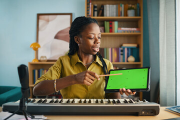 African American woman holding tablet with green screen while teaching online piano lesson at home. Room filled with books and modern decor creating an engaging learning environment
