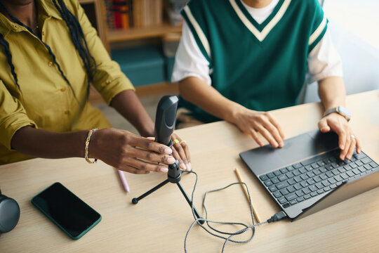 Two colleagues setting up a microphone for podcasting with an open laptop on desk. Various digital gadgets placed on table creating functional workspace