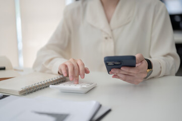 Female office worker holding a phone and using calculator while sitting at working table in office.