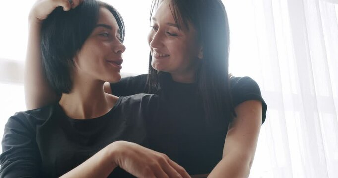 Couple, lgbt and lesbian women at home. Low angle handheld shot of young lesbian couple smiling and touching hands gently while sitting on sofa against window in light living room at home.