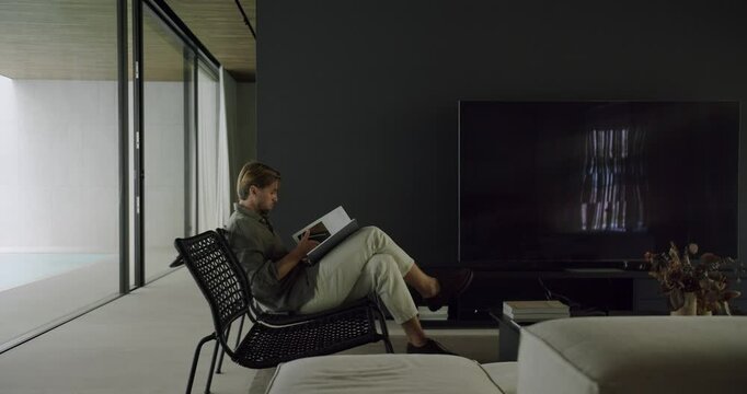 A man relaxes in a wicker chair by a large window, immersed in a book within his sleek, modern living room. Man Relaxing in Modern Home with Book by Large Window