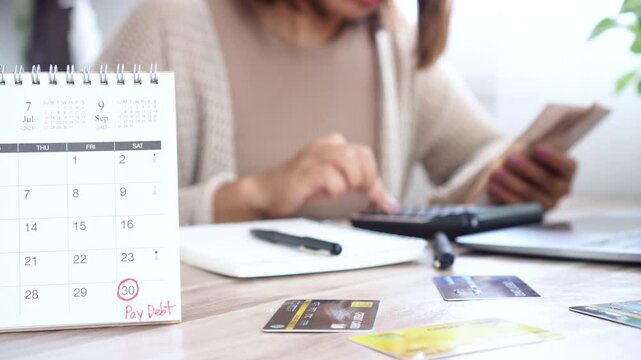 Close-up calendar with a reminder note to pay debt by the end of the month, as a woman counts money in her hand, checks income and spending, and uses a calculator with credit cards on the table 