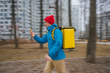 Courier wearing a yellow insulated backpack and red beanie, running through an urban park while...