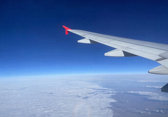 Obraz premium Airplane wing above the clouds. A view from a plane window showing the aircraft wing high above a thick layer of clouds. The deep blue gradient sky transitions into the hazy horizon.