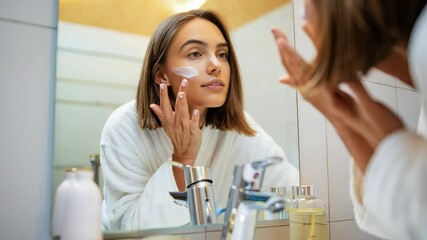 Young woman in a bathrobe applying moisturizing cream to her face while gazing into the bathroom mirror, embracing her morning skincare routine with a sense of relaxation and self-care - Powered by Adobe