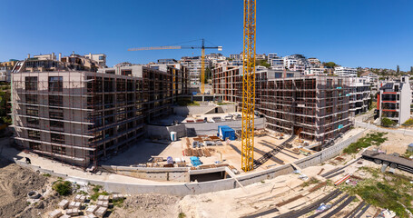 Panoramic view of a modern residential construction site with scaffolding, cranes, and unfinished buildings under a clear blue sky in an urban environment