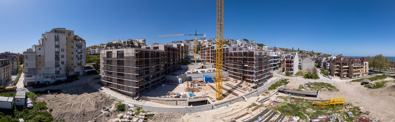 Panoramic view of a modern residential construction site with scaffolding, cranes, and unfinished buildings under a clear blue sky in an urban environment