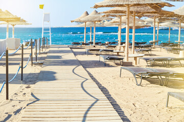 Fototapeta premium Beach umbrellas parasol made of wood and sunbeds standing on seashore against the backdrop of sunny blue sky and glare. View over tropical beach with rows of parasols. Vacation and Travel Concept.