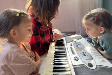 Teen boy shows his twin toddler sisters how to use a synthesizer. The kids sit on the floor, laughing and learning through music. Happy family bonding indoors, real life