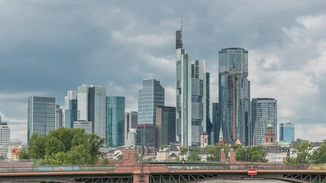 Aerial hyperlapse of Frankfurt skyline with Main River and Ignatz Bubis Bridge. Skyscrapers and church spires reflected in the water beneath dramatic clouds timelapse. Frankfurt, Germany