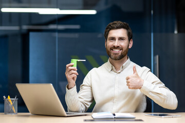 Portrait of a young smiling man sitting in the office at his desk, holding an aerosol spray can and...