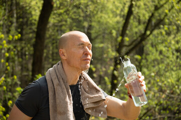 Sporty man refreshing and splash himself water from bottle against forest at summer heat after...
