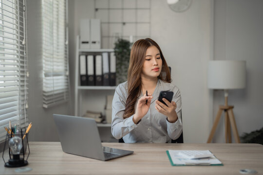 Young Asian businesswoman holding pen and using smartphone while working with laptop computer at office desk, business and technology concept - Powered by Adobe