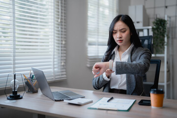 Young Asian businesswoman looking at her smartwatch while sitting at desk in modern office, waiting...