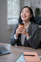 Asian businesswoman sitting at her desk, enjoying a coffee break with closed eyes and smiling, while working on a laptop and taking notes in a notebook
