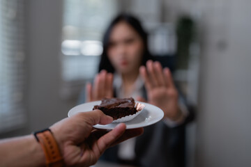 Businesswoman is raising her hands and refusing a chocolate brownie offered by a colleague, showing...