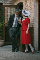 Handsome brutal man and beautiful woman stand close beside wall, dressed old-fashioned attire, with drinks in hand, sharing calm talk. Vintage style and gentle mood. Concept of vintage, love.