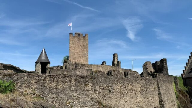 Medieval Bourscheid Castle. Stone walls and watchtowers. Flag is waving on top. Modern wind turbine spins on the hill in the background. Bourscheid Castle, Luxembourg 