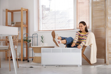 Young woman sitting on armchair near electric heater in living room