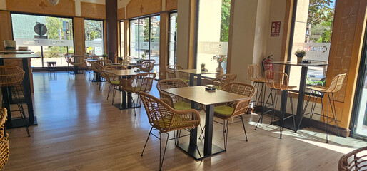 Modern cafe interior with wicker chairs, square wooden tables, large glass windows, high bar tables, and natural light on a wooden floor; Spanish greenhouses;
