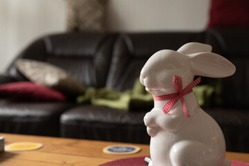 White ceramic bunny with red bow, sitting on a coffee table in a living room. Spring, Easter, home decor.