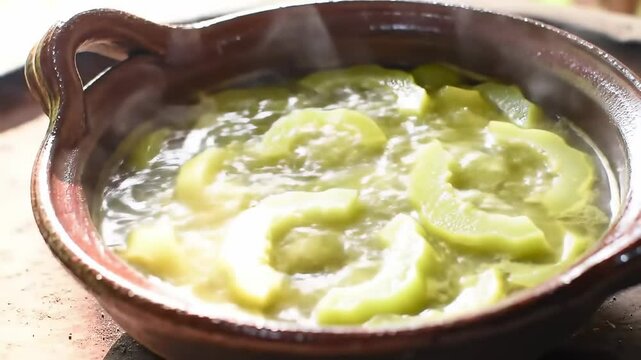 A traditional clay pot filled with simmering green squash slices in a rustic kitchen setting - sayote | chayote