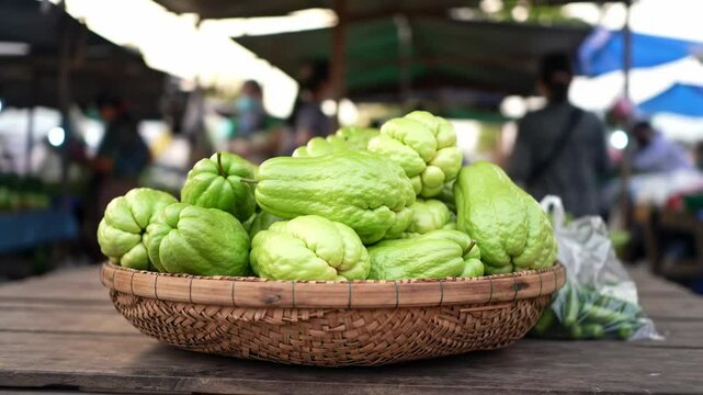 Fresh green chayote squash piled in a woven basket at a bustling market with shoppers in the background - sayote | chayote