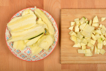 Peeled and chopped zucchini pieces are arranged on a patterned plate and a wooden cutting board, ready for cooking