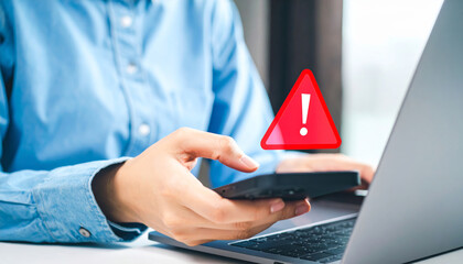 Person holding smartphone with warning alert symbol on screen, sitting at desk with laptop, concept of cybersecurity threat, data breach, or ransomware attack, feeling worried and cautious