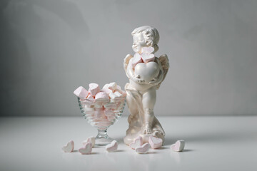 Valentine's Day composition featuring a cupid figurine and a glass filled with pink marshmallows shaped like hearts. Minimalist and dreamy setup.