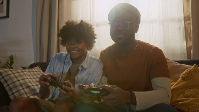 Tilt up shot of African American father and son gently punching each other with laughter during friendly competition in exciting video game session, sitting on couch in sunlit apartment