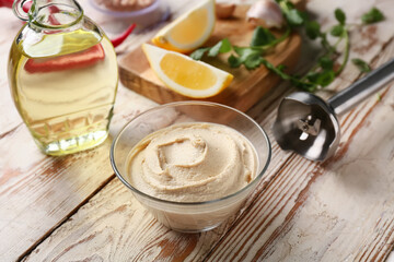 Bowl of tasty hummus with ingredients and blender on light wooden background, closeup