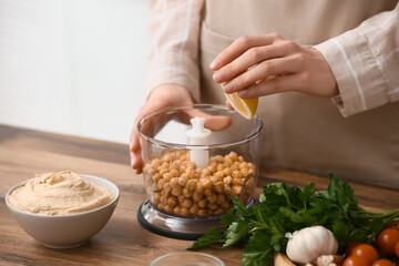Young woman adding lemon juice into blender with chickpeas for hummus on table in kitchen